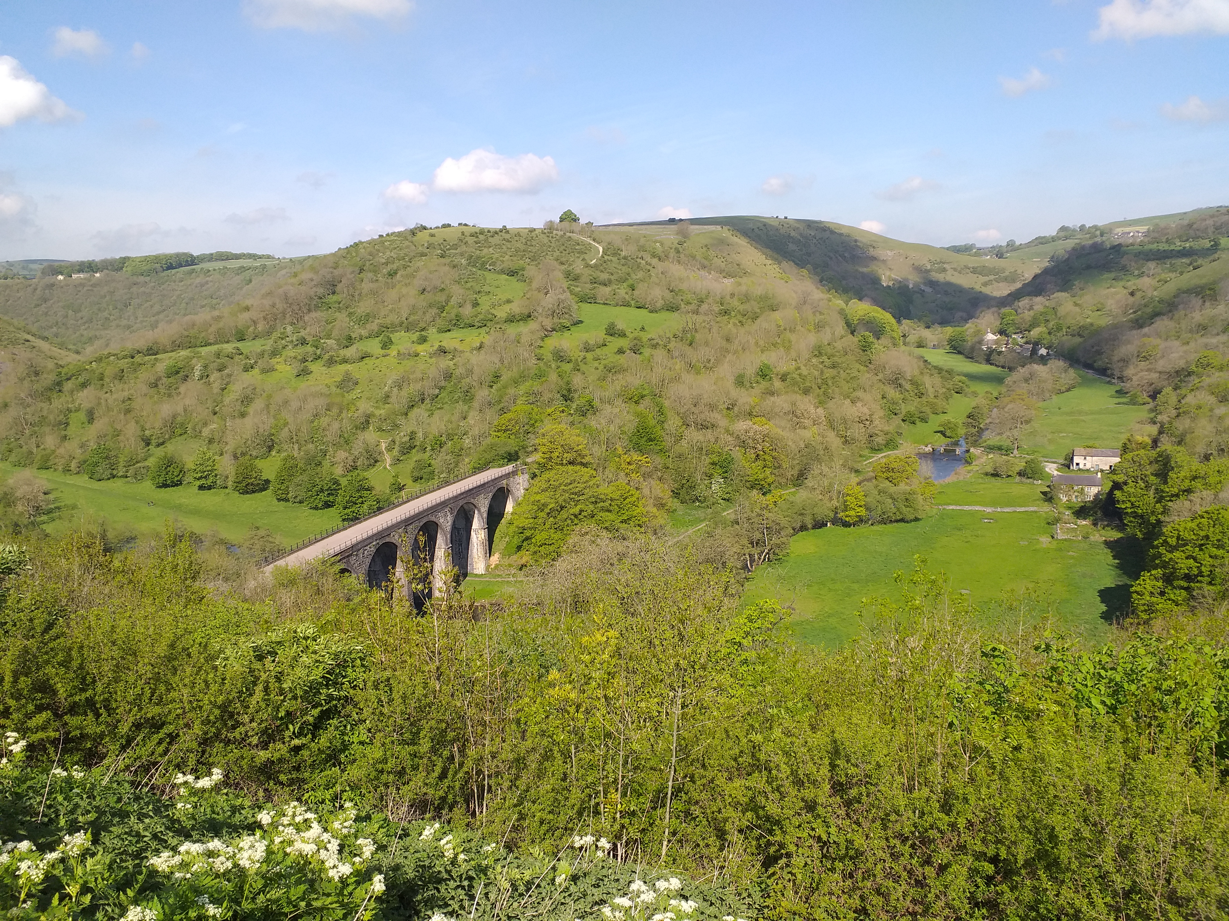 As view over the Monsal Valley with rolling green hills and the stone railway viaduct in the centre, blue skies