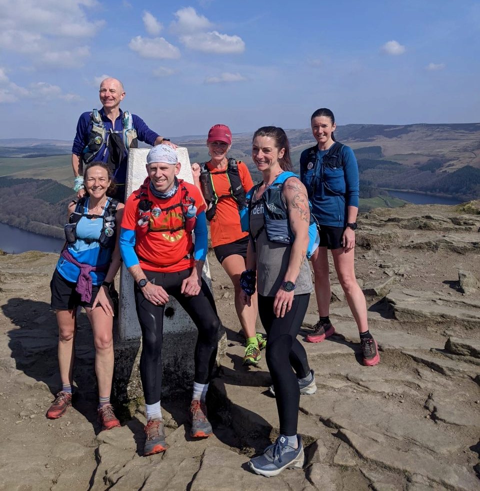 A group of smiling runners stand next to a white Peak District trigg point