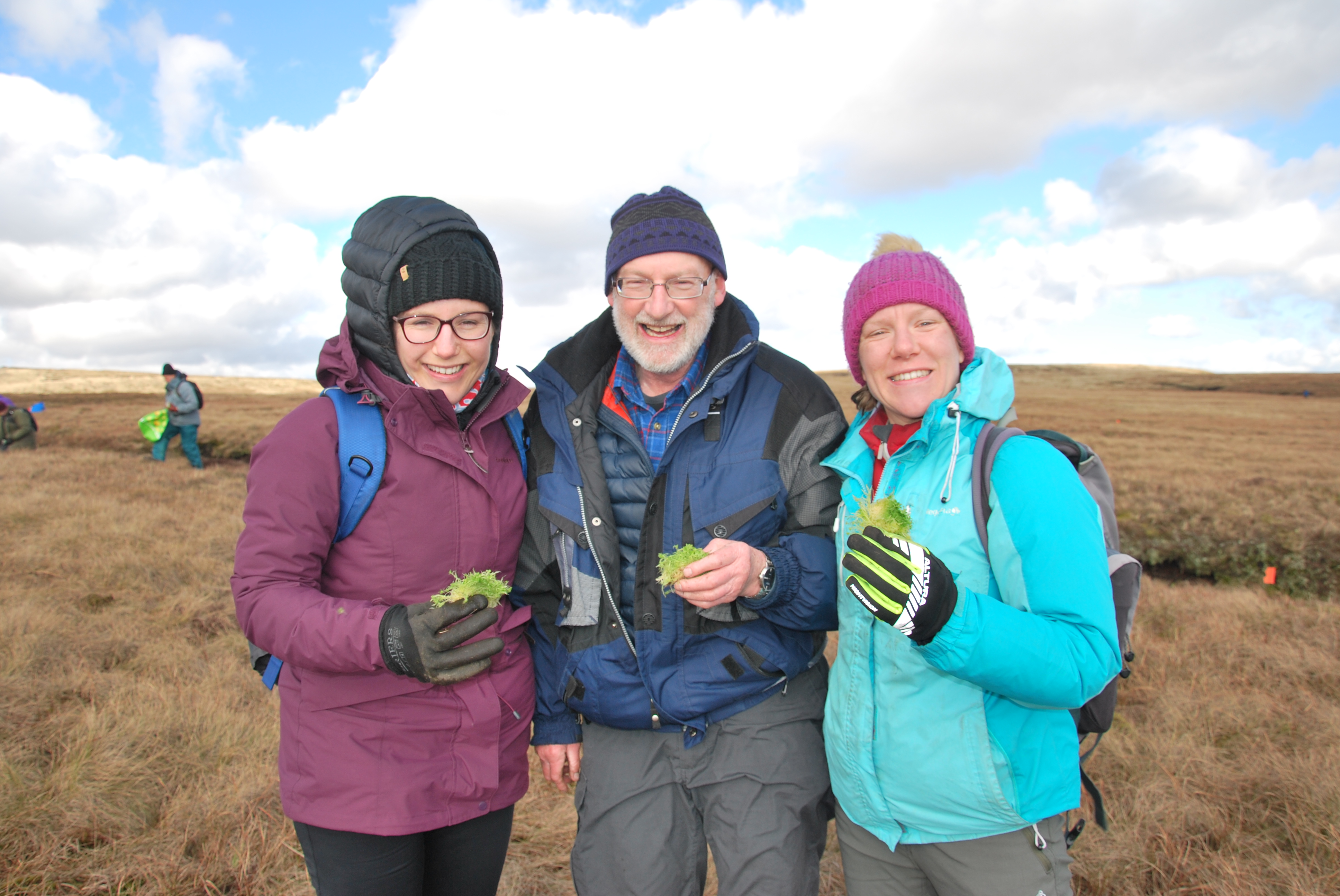 Three volunteers pose in outdoor clothing with a Peak District moorland landscape behind them
