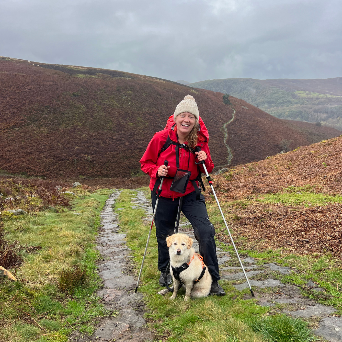 Jen stands with walking poles on a hill in the Peak District with her cream-coloured dog Cookie. It is a cloudy and rainy day. She wears a red coat and a bobble hat.