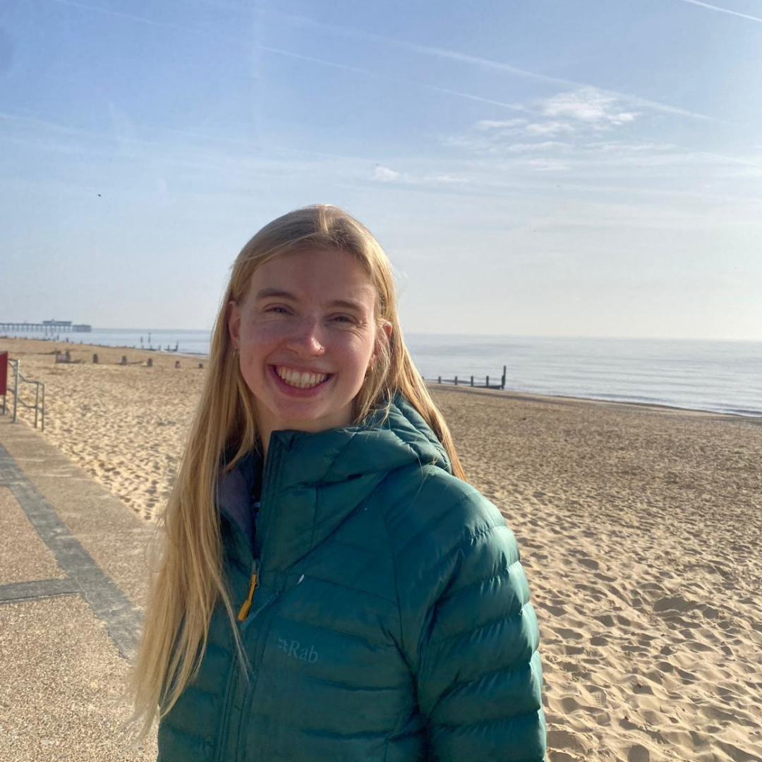A blonde woman in a coat stands by the beach.