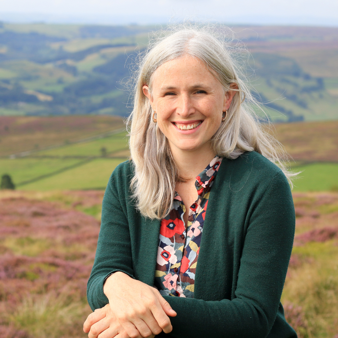 A blonde woman sits in front of a Peak District scene.